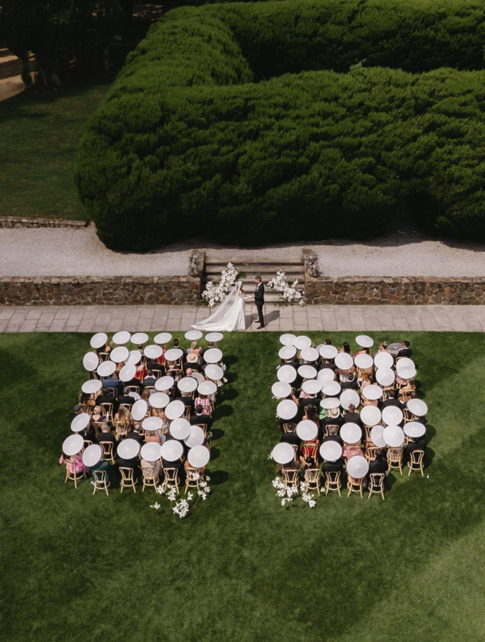 An aerial view of a garden ceremony: guests under white parasols either side of the aisle, the couple at lily-lined stone steps