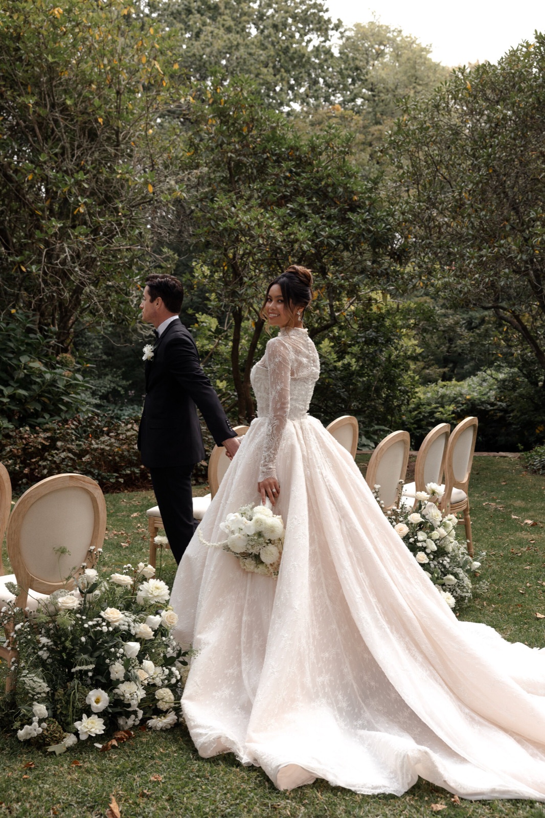 A bride in a long-sleeved gown walking to her groom past flower-lined chairs