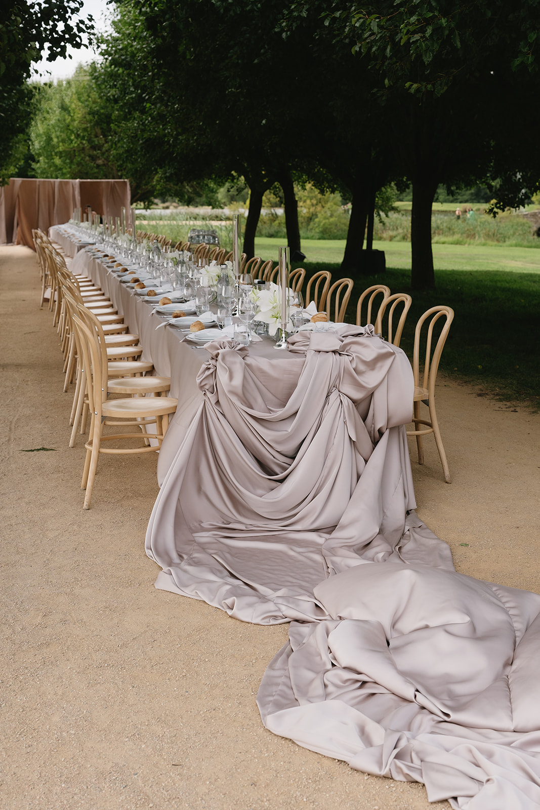 A long reception table under trees, its blush satin cloth gathered and pooling onto the gravel path