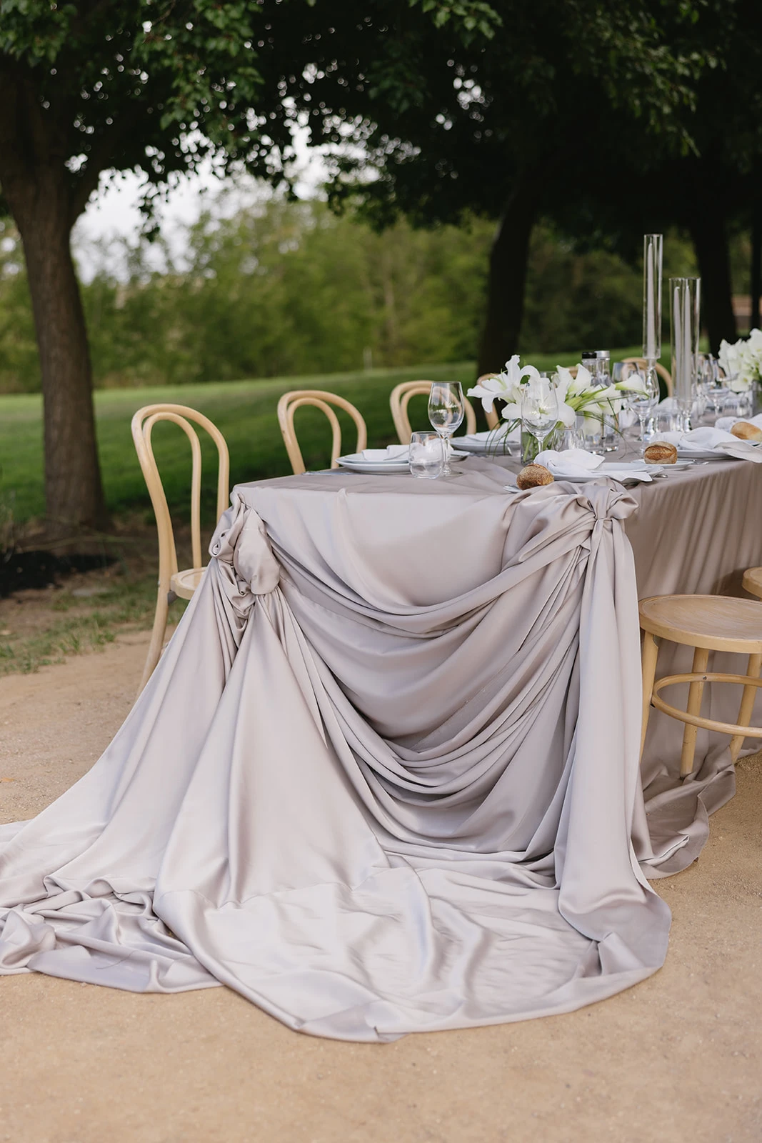 A close view of a table end wrapped in gathered blush satin that pools onto the gravel, white lilies and candles behind