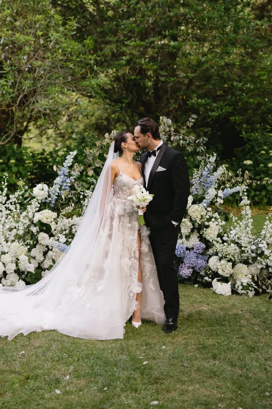 A bride and groom kissing in front of a ceremony arch of white and blue flowers in a garden
