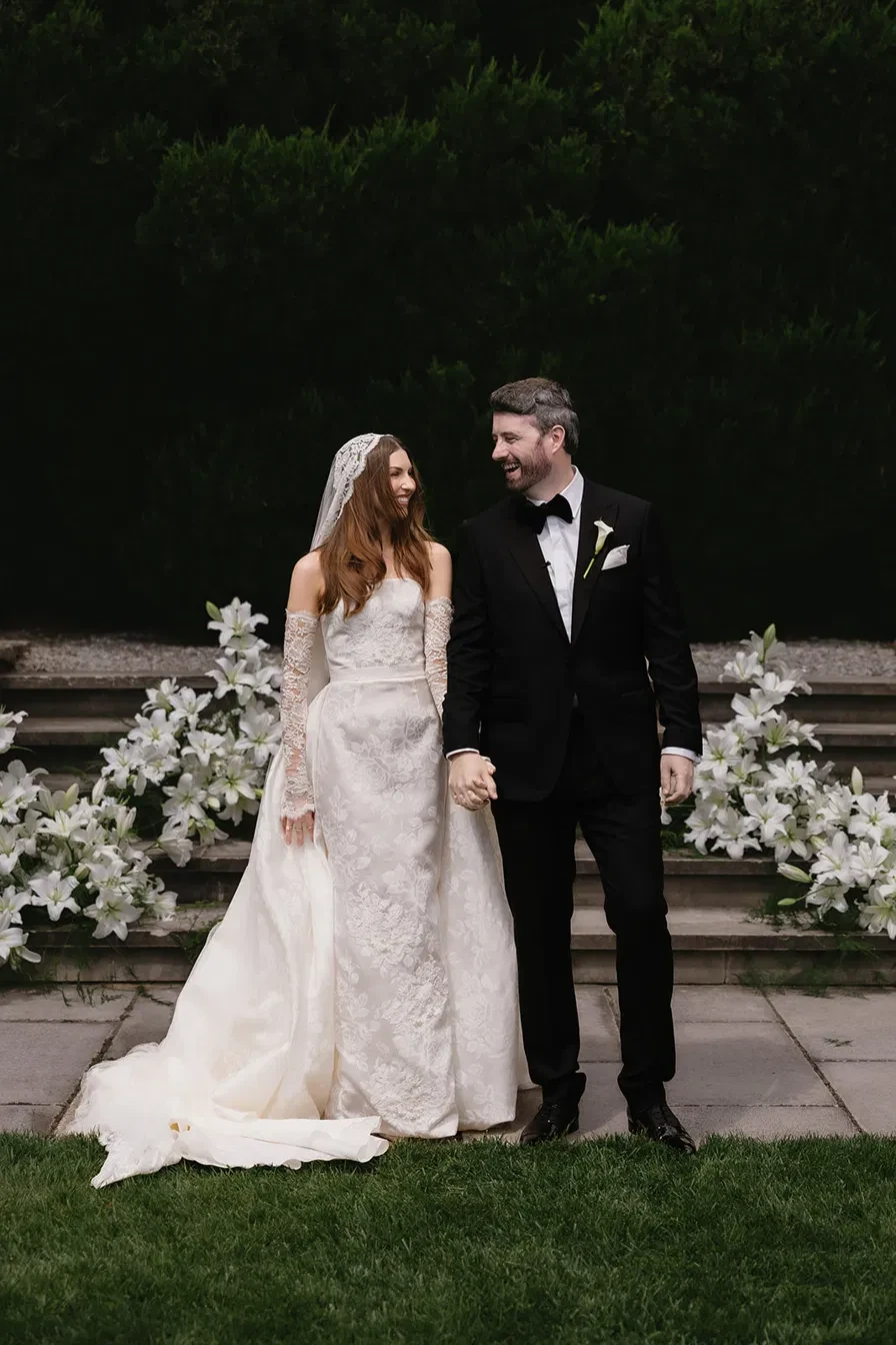 A bride and groom holding hands and laughing in front of stone steps lined with white lilies