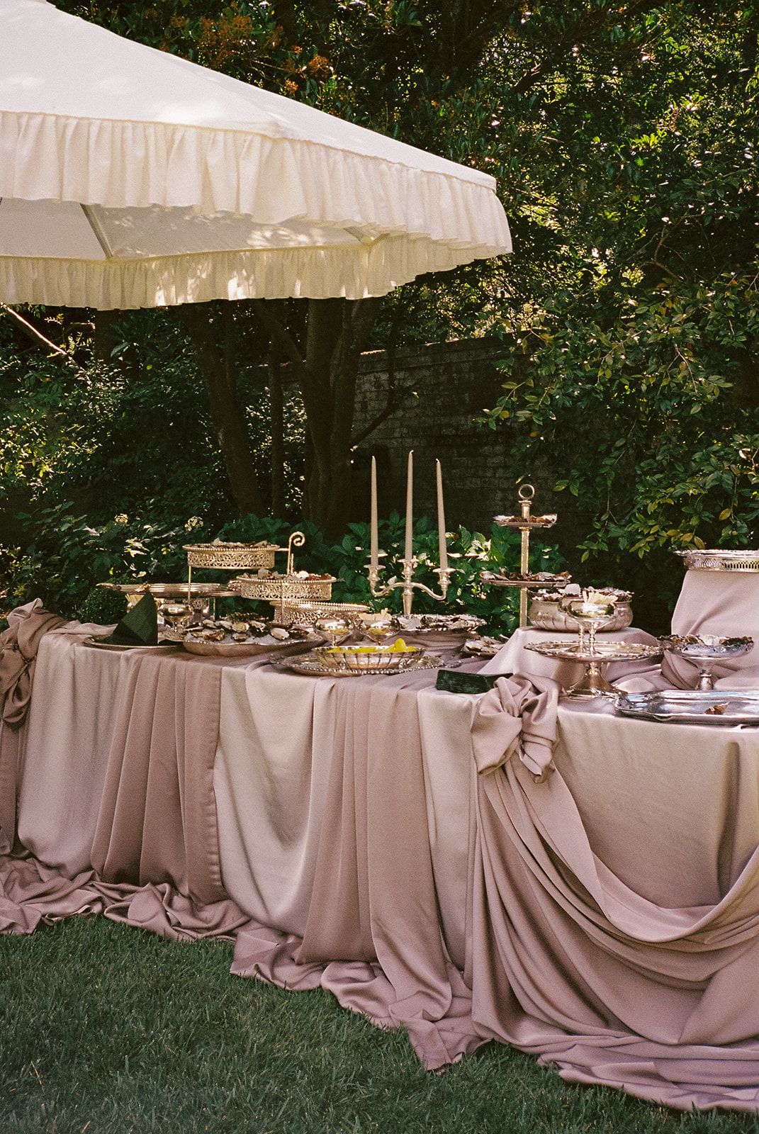 An oyster and champagne table draped in blush satin under a ruffled white umbrella in a garden
