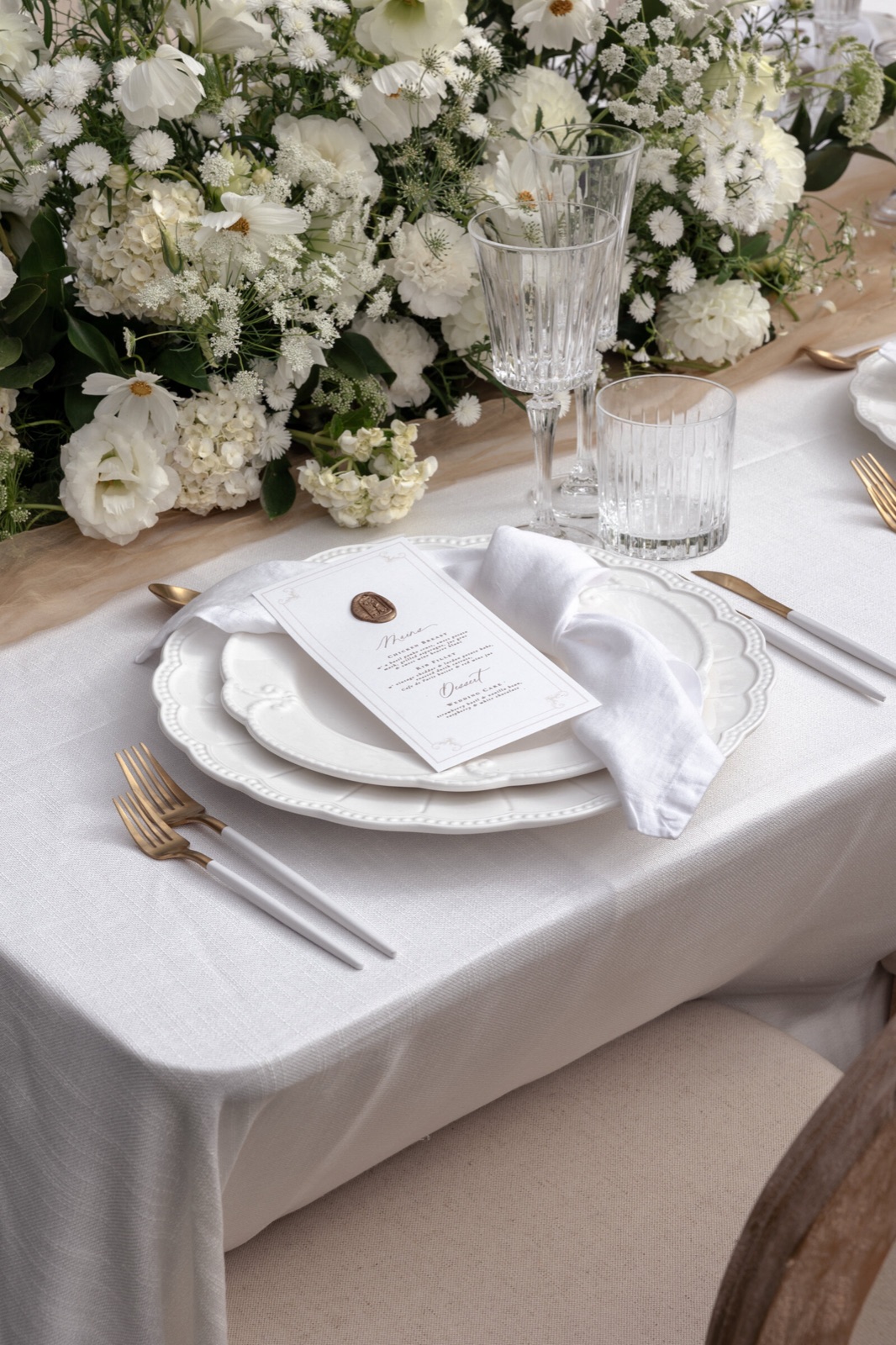 A table set with crystal glasses, a wax-sealed menu and a white napkin under white flowers